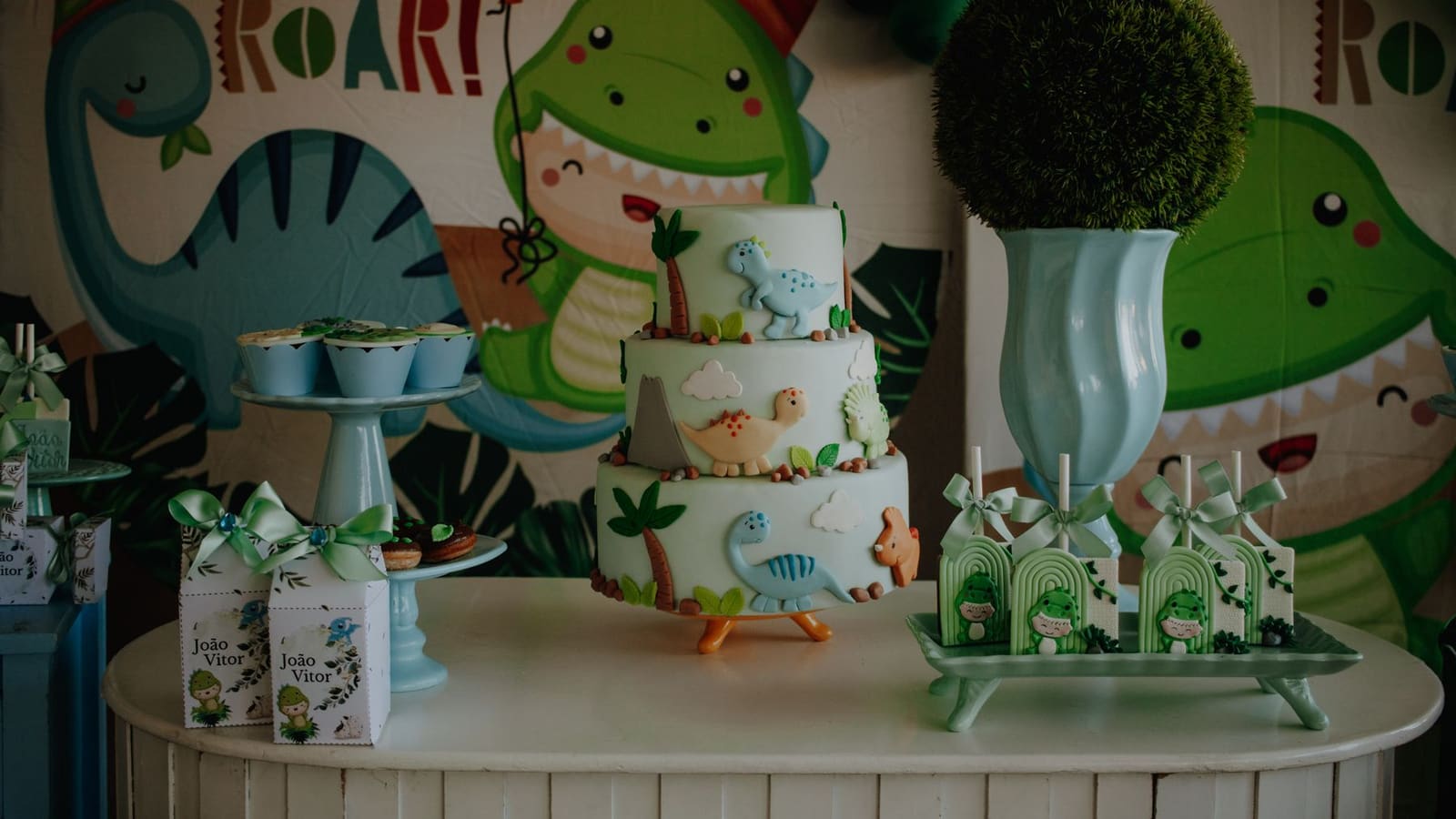 A dinosaur-themed birthday dessert table with a three-tier dinosaur cake and a 'ROAR!' backdrop