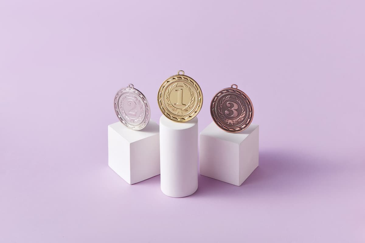 Gold, silver, and bronze medals displayed on white pedestals with a pink background