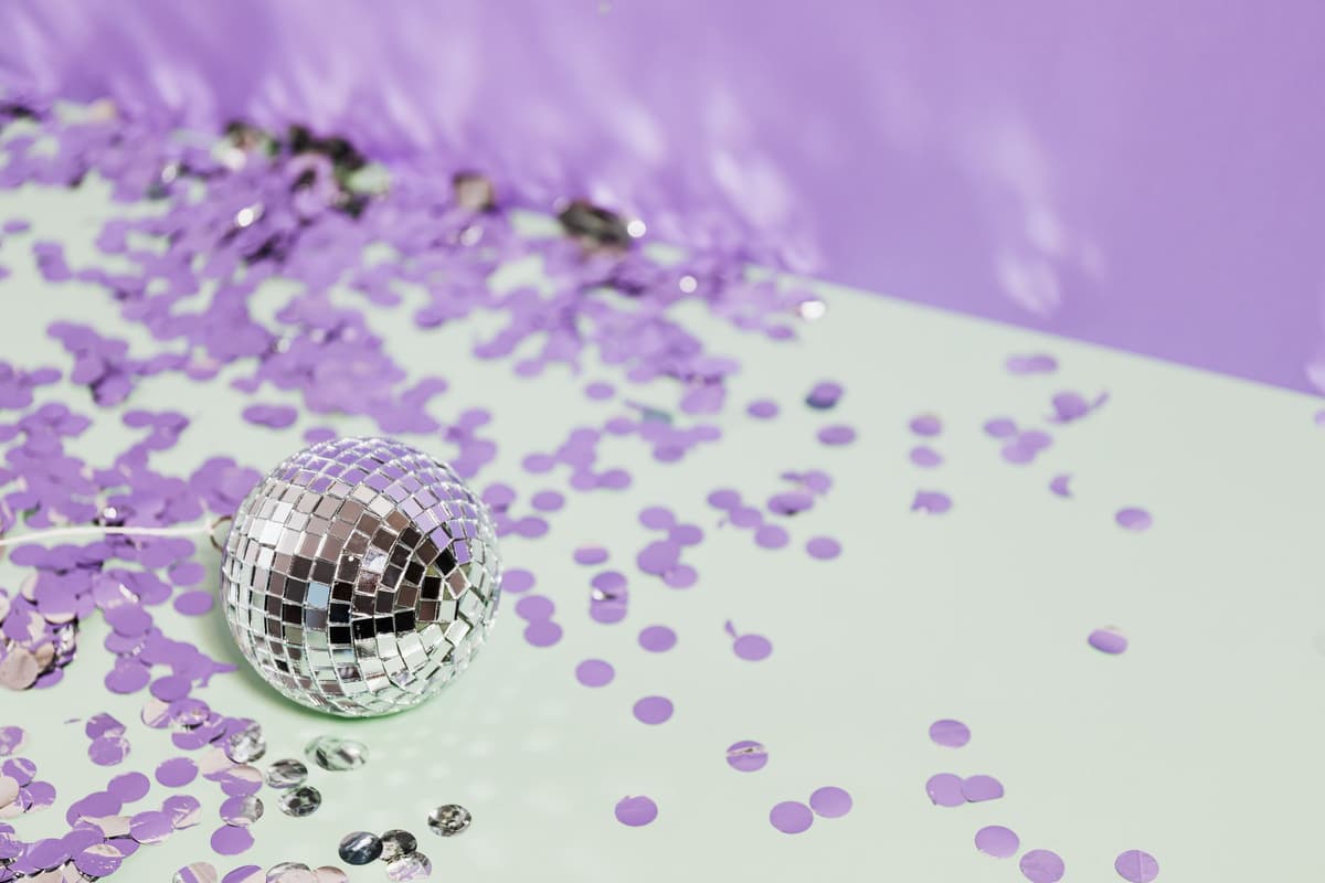 A shiny disco ball surrounded by purple confetti on a pastel background, evoking a festive mood