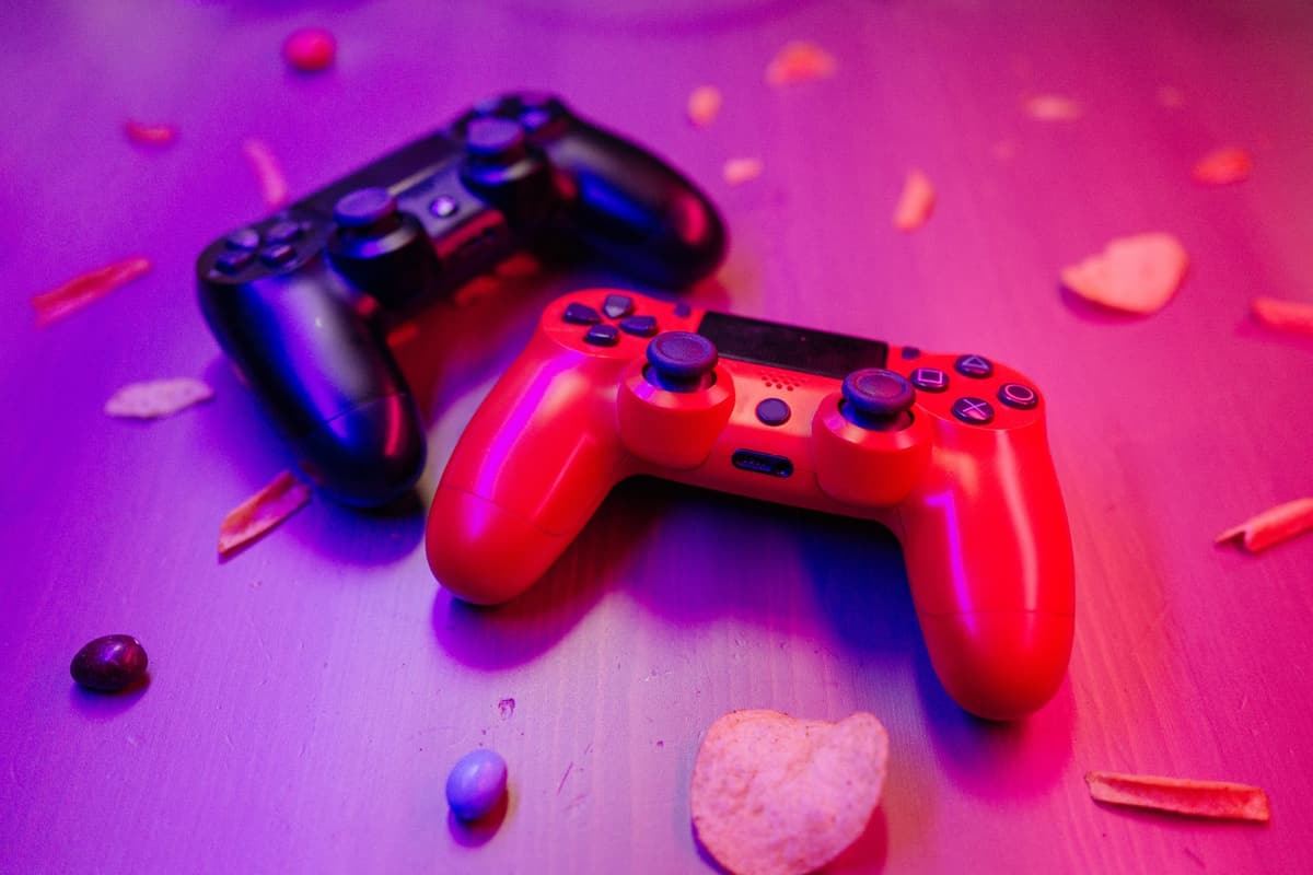 Close-up of red and black game controllers on a table with snacks around, lit by ambient colored light
