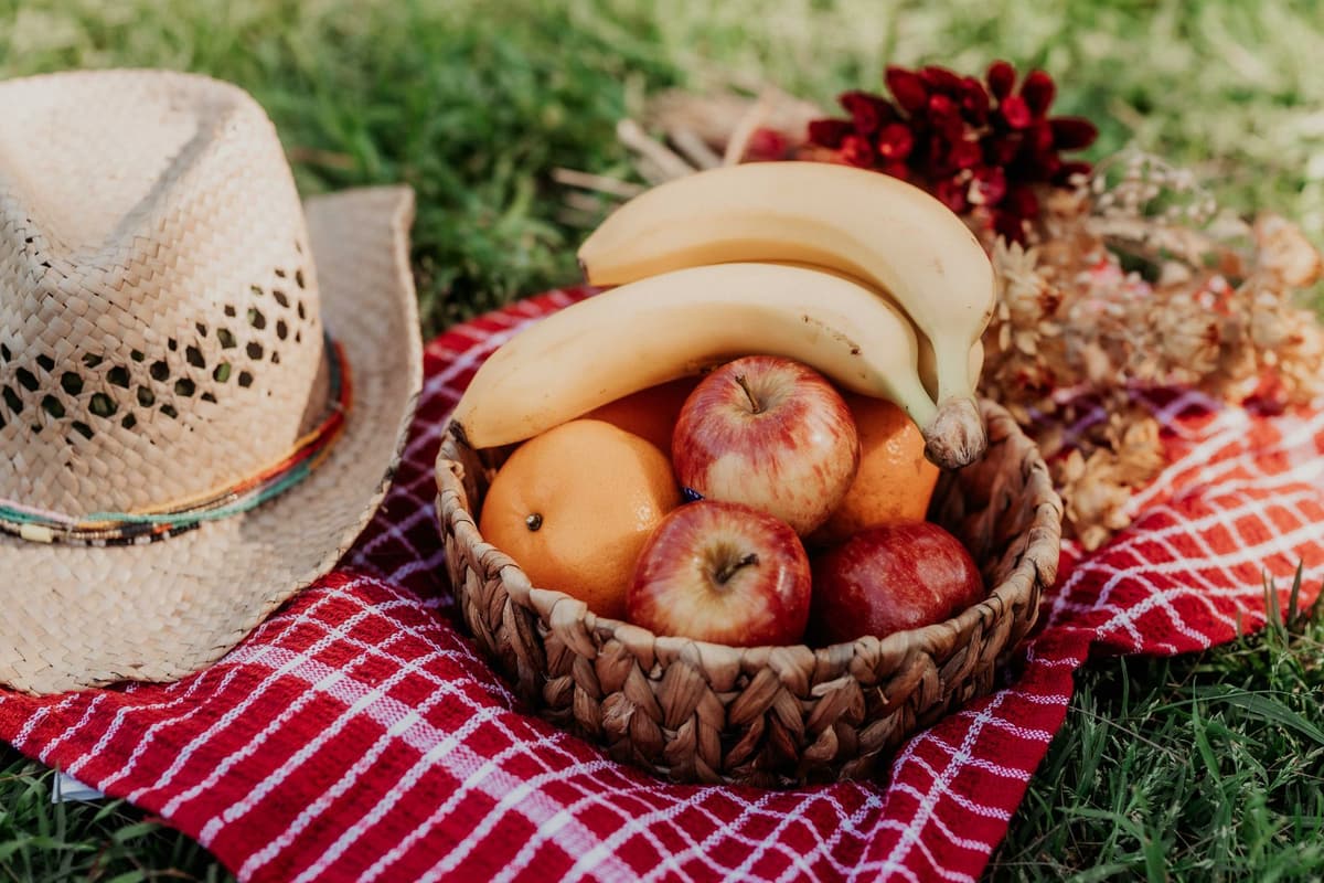 Vibrant picnic setting with a straw hat and fruit basket on a red checkered cloth, perfect for outdoor leisure
